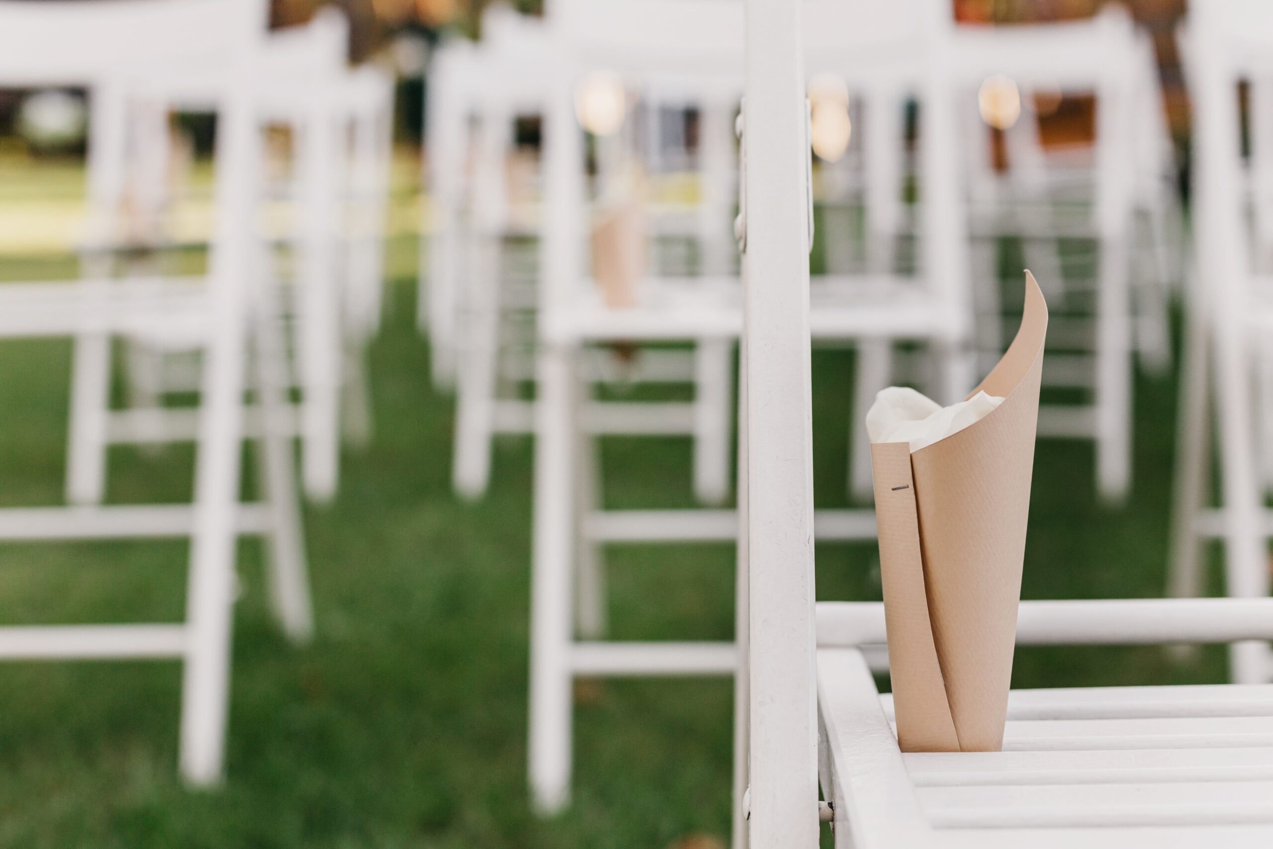 White folding chairs arranged on a green lawn with paper packages filled with petals, prepared for an outdoor marriage ceremony.