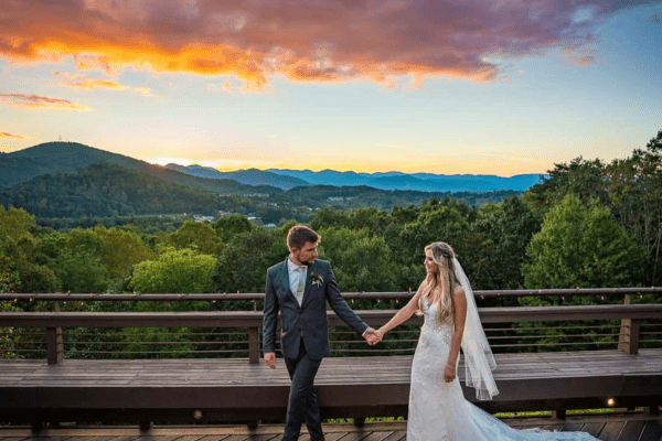 A bride and groom hold hands on a scenic deck in Greenville, NC, with a breathtaking mountain sunset in the background.